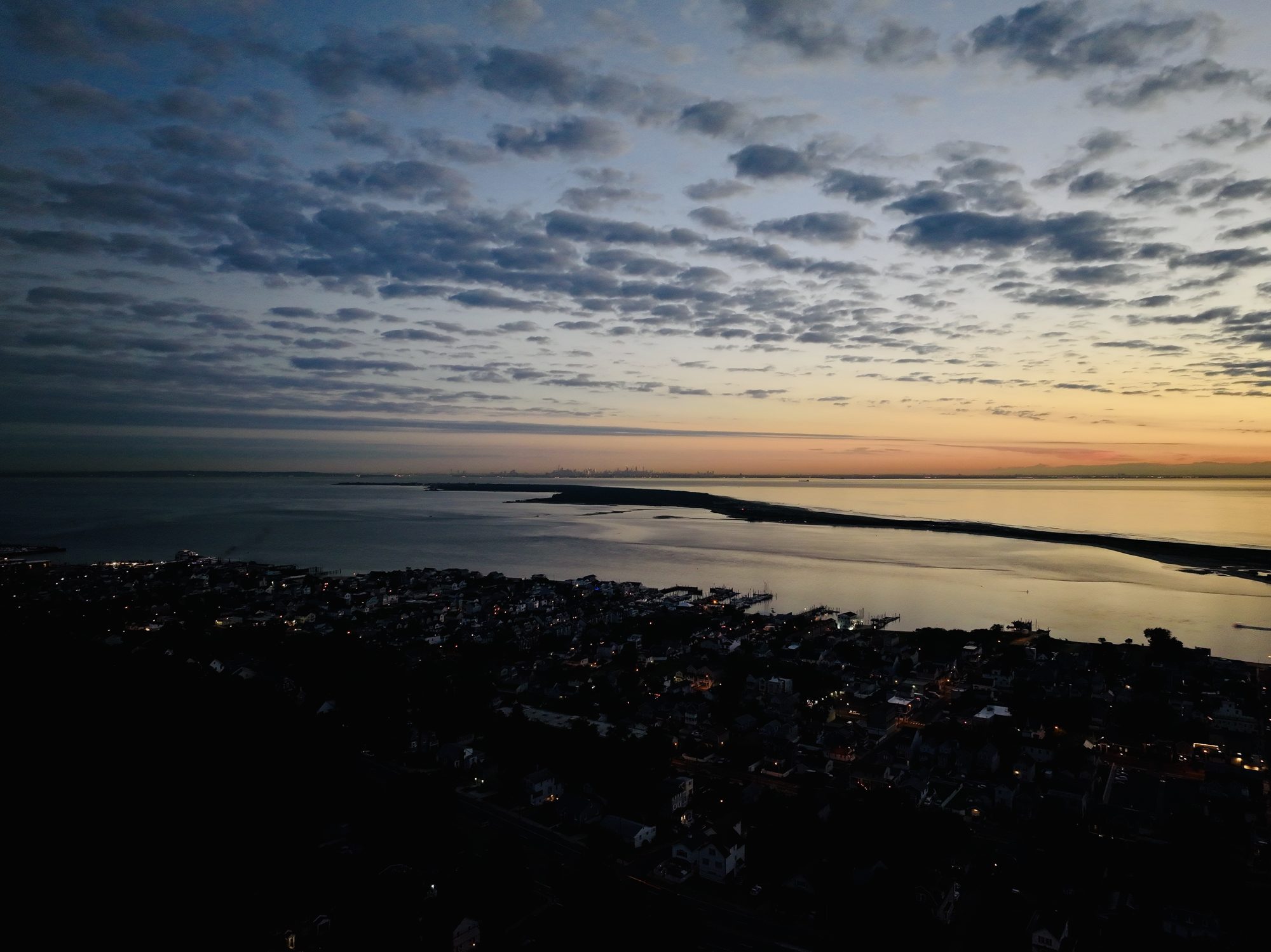 Aerial photograph at dawn of Highlands, New Jersey — town lights on the hillside, Sandy Hook Bay, the barrier spit reaching north, and the Manhattan skyline faint on the horizon, 2026