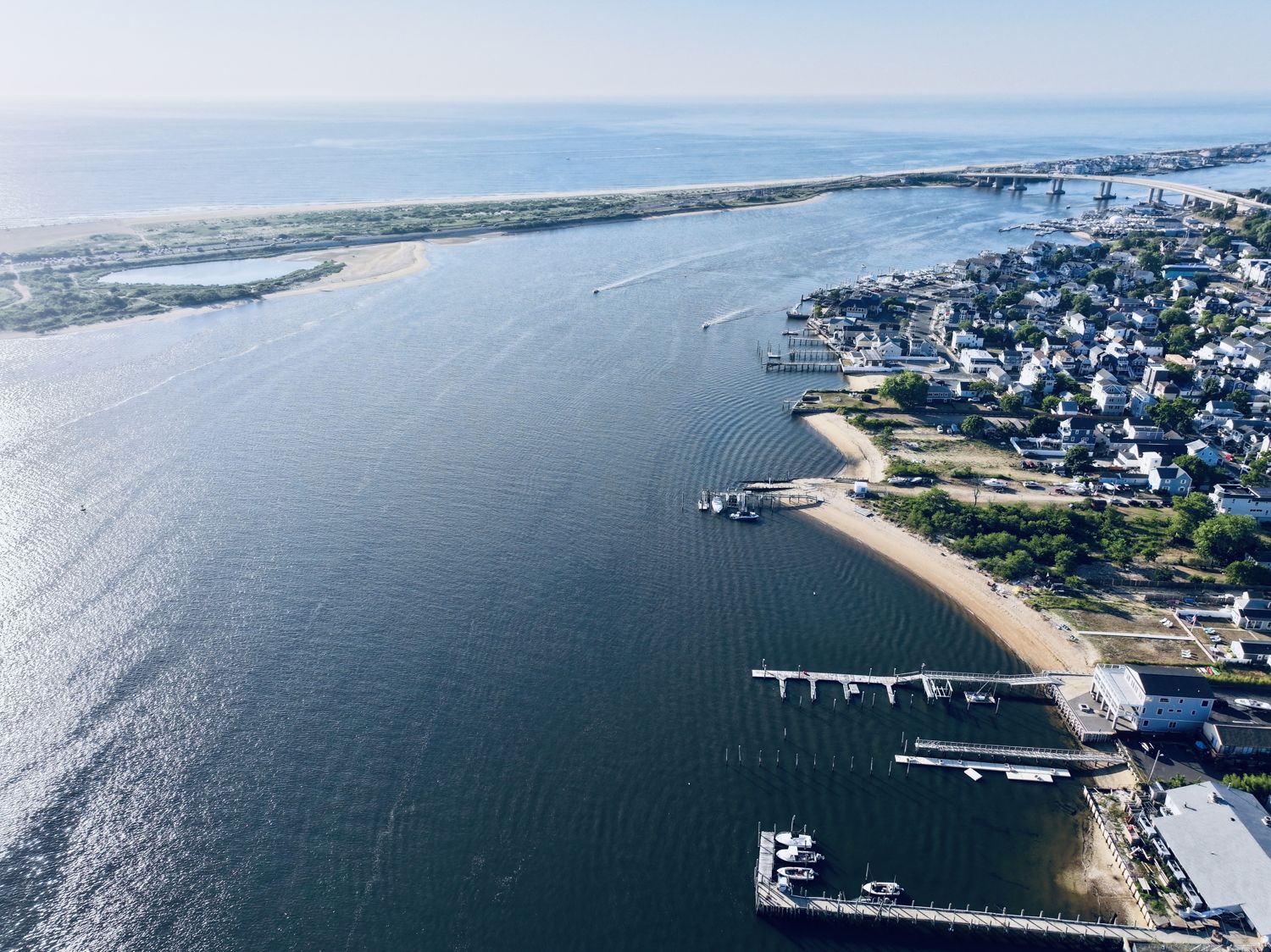 Aerial photograph of the mouth of the Navesink River opening toward Sandy Hook, houses along the far shore and a pier reaching into the channel, 2026
