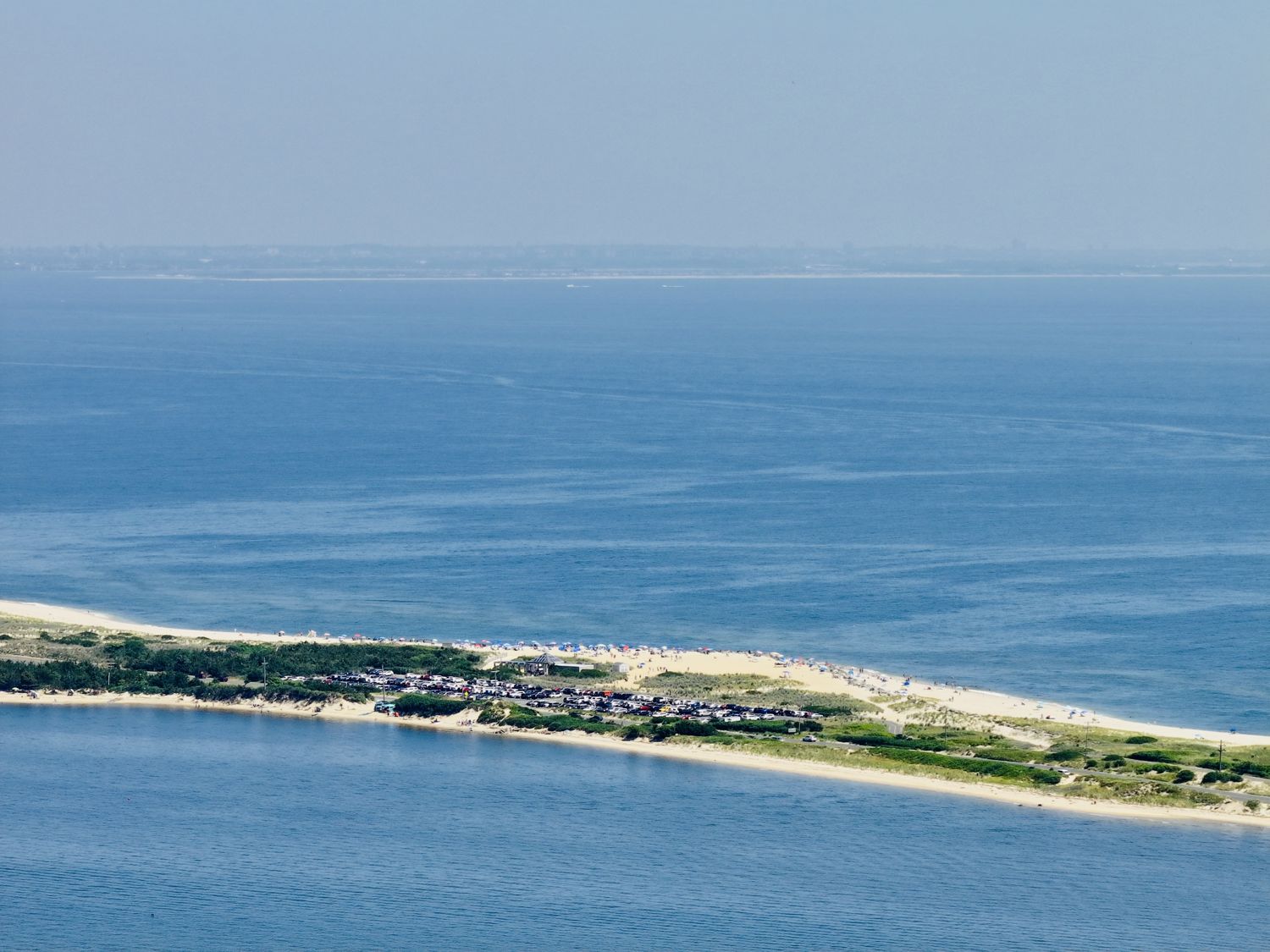 Aerial photograph of Sandy Hook from the north, the wooded spit and public beach in the foreground, the towers of Manhattan on the horizon, 2026