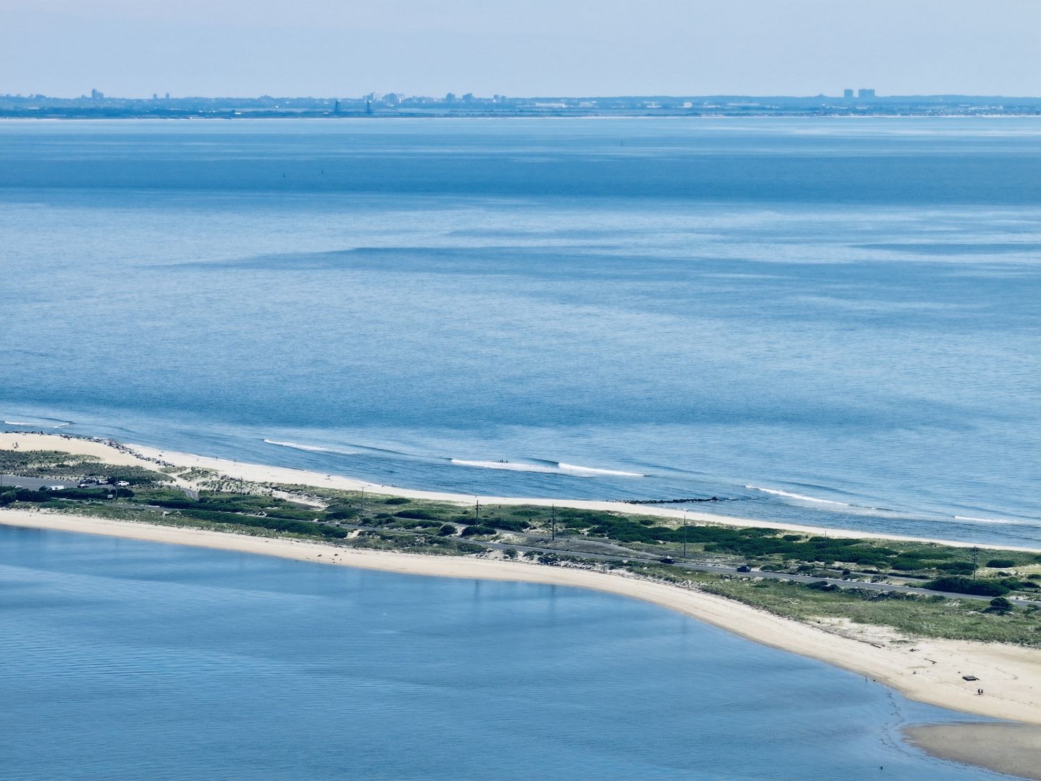 Aerial photograph of the Sandy Hook barrier spit curling north into Lower New York Bay, ocean on the right, bay on the left, the Manhattan skyline faint on the horizon, 2026