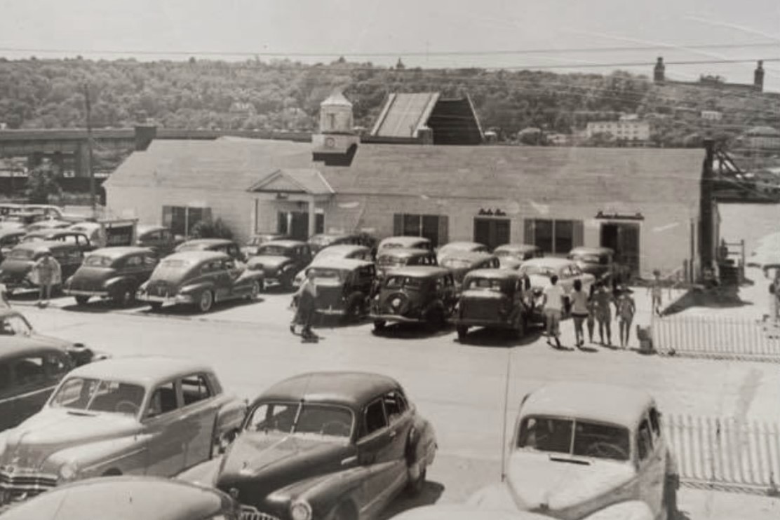 The Sandlass Beach Club at Highland Beach on a summer Saturday ca. 1948 — the Bamboo Room building with parking lot full of 1940s American sedans, families walking toward the beach, the Highlands ridge behind