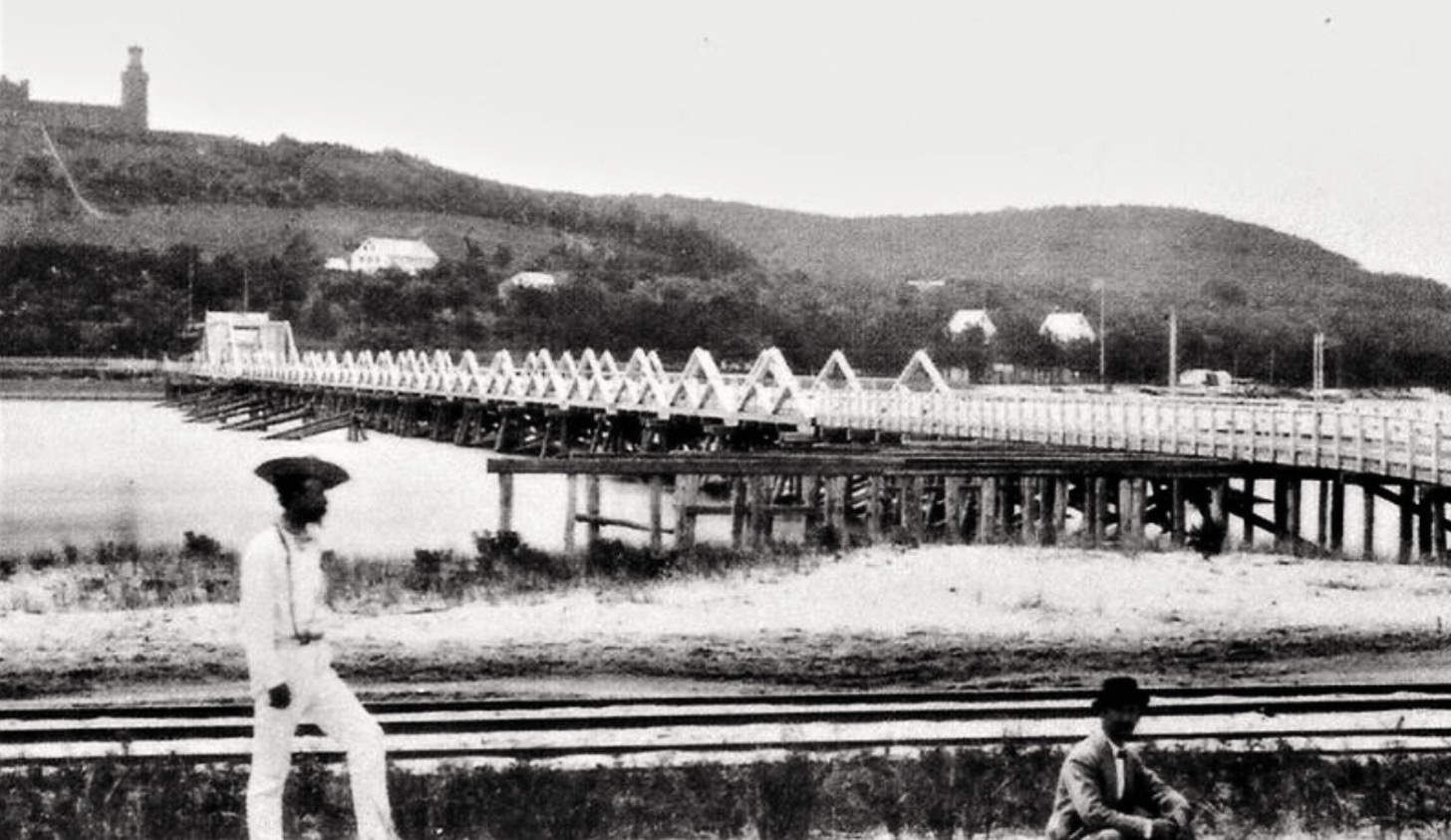 A black-and-white photograph from the ferry pier at Highland Beach ca. 1900 — the sawtooth wooden Sandy Hook bridge crossing the Shrewsbury, Twin Lights on the ridge, two figures in period dress