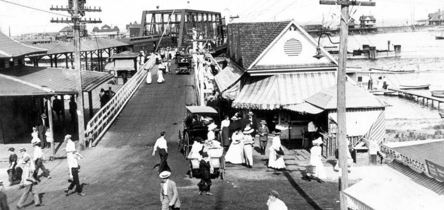 The Highland Beach ferry pier ca. 1905 — pedestrians in Edwardian dress arriving from the covered station platforms, a horse-drawn wagon on the pier, the iron-truss Shrewsbury bridge crossing behind, a Cigars and Tobaccos stall at right
