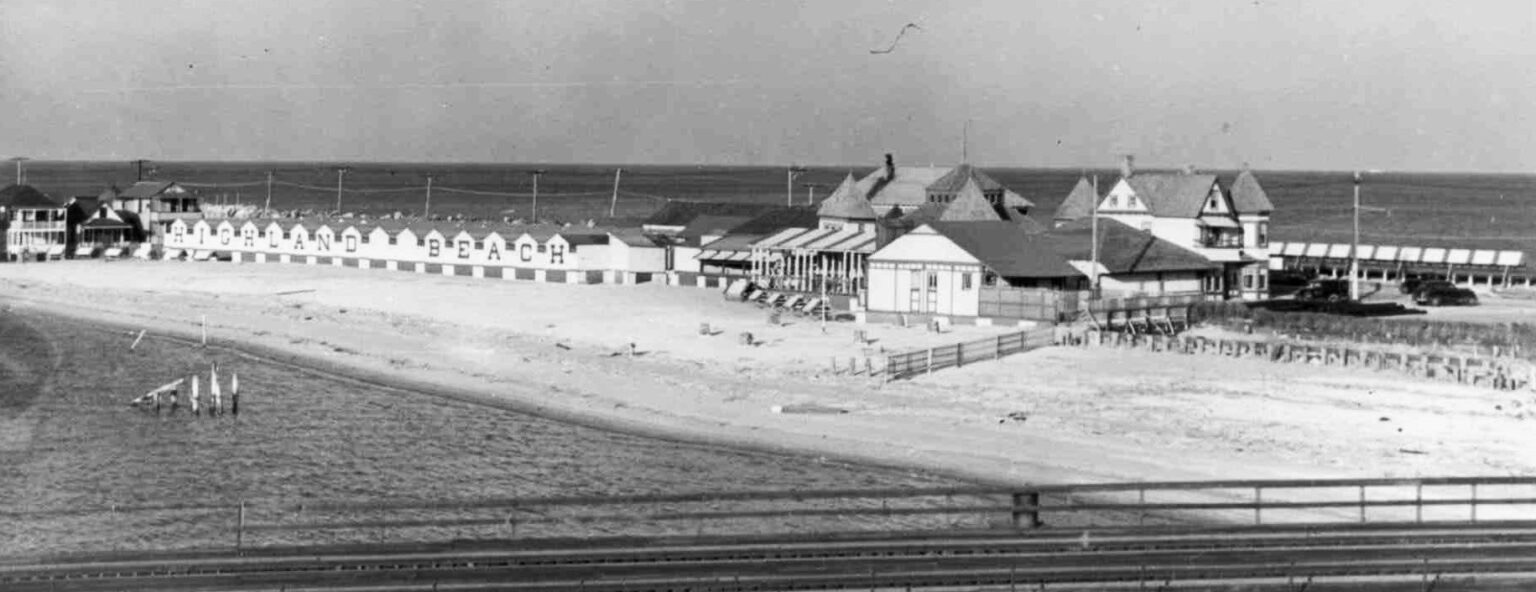 The Sandlass Beach bathhouse row at Highland Beach, ca. 1925 — the long low structure with HIGHLAND BEACH painted in white block letters across its seaward face, the cottage colony at left, the Basket Pavilion at right