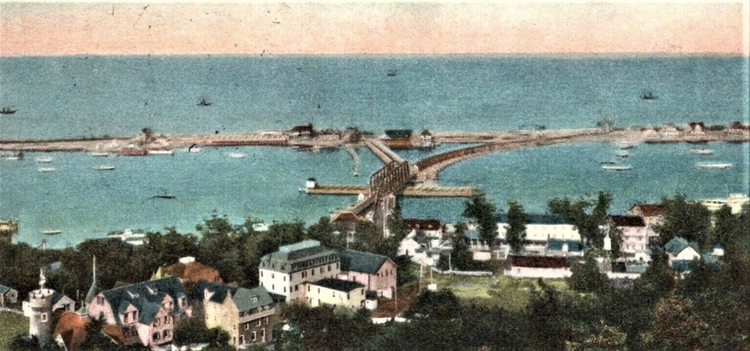 A hand-colored postcard aerial view ca. 1910 — from the Highlands bluff across the Shrewsbury river to the Sandy Hook bay, with the sawtooth iron-truss bridge, sailing craft in the bay, and the pale Sandy Hook spit on the horizon