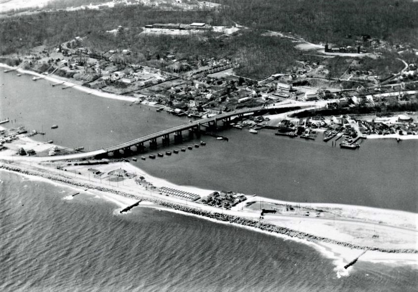 An aerial photograph ca. 1948 looking east — the Route 36 high bridge crossing to Sandy Hook, the Sandlass Beach Club bathhouses and family house at the foot of the bridge, small craft in the docks