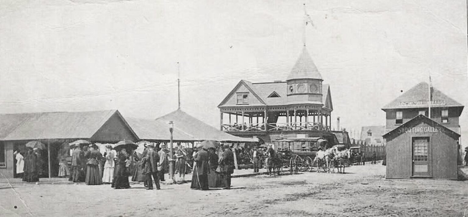 A sepia photograph ca. 1898 of the Basket Pavilion at Highland Beach — three-story wooden dance hall with hexagonal tower, station platform at left with passengers in Edwardian dress, horse carriages in the middle, and a SHOOTING GALLERY building at right