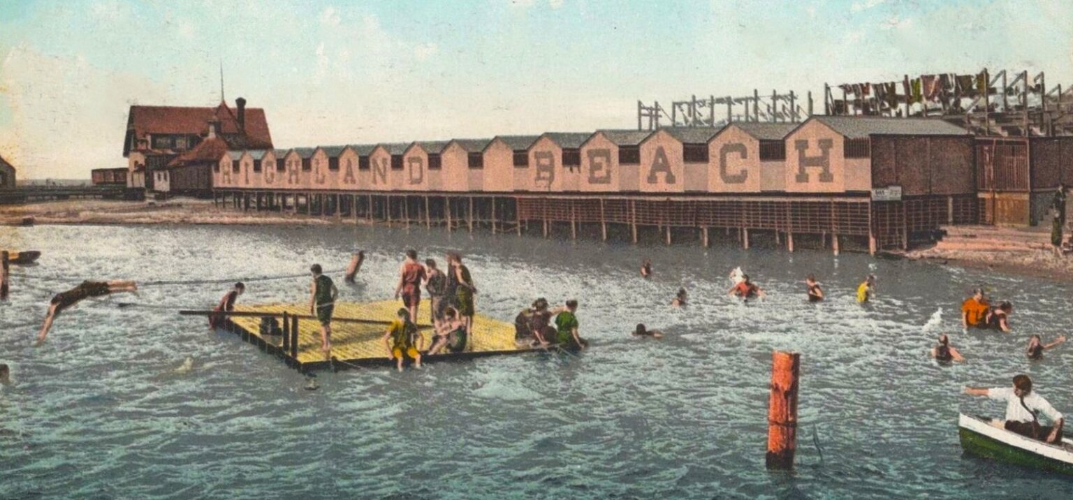 A hand-colored postcard ca. 1908 of the Highland Beach bathhouse row on wooden pilings with HIGHLAND BEACH painted across its face, a wooden floating raft in the swimming area, bathers, and the roller-coaster trestle behind