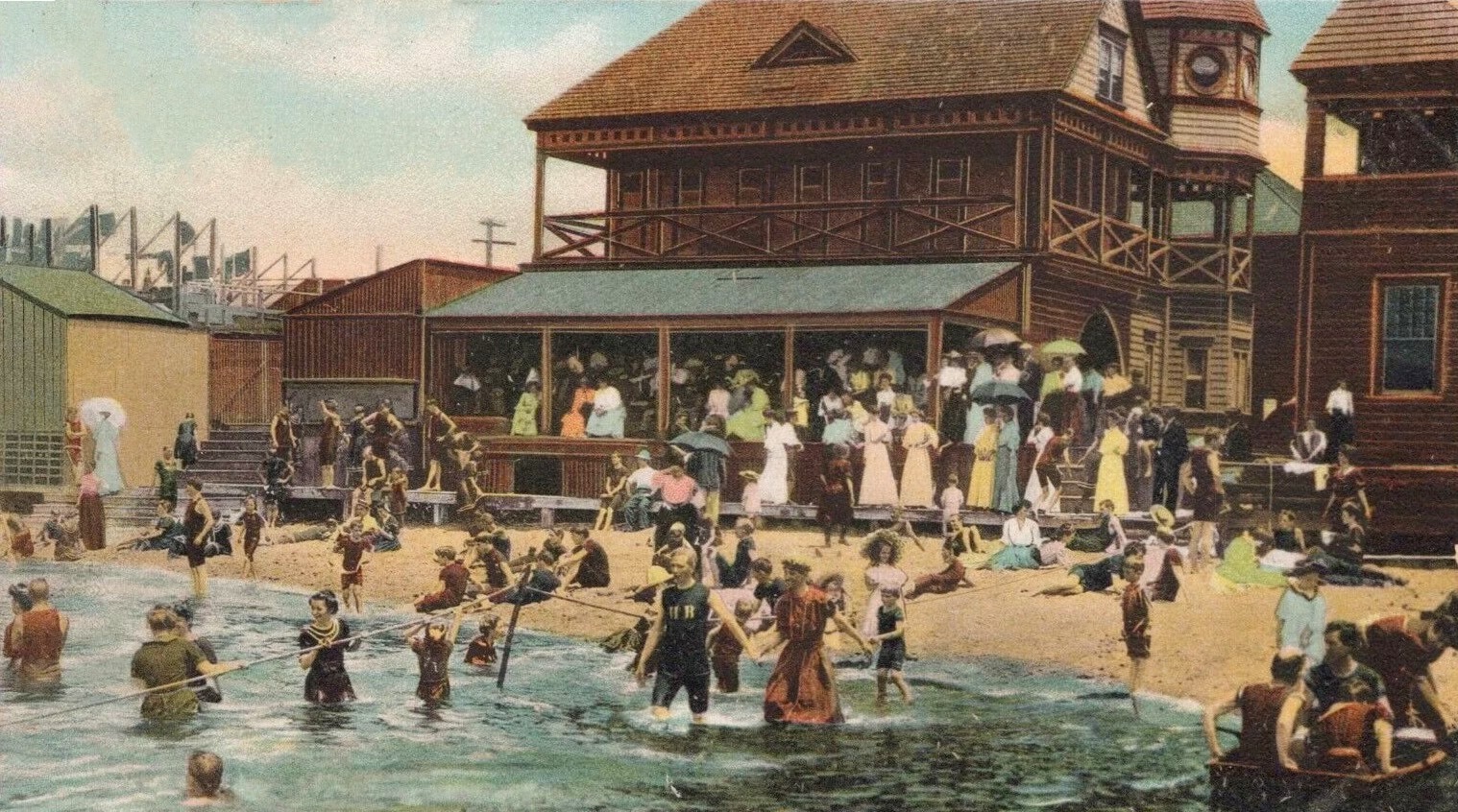 A colorized postcard of the Sandlass Pavilion at bathing hour ca. 1908 — the Basket Pavilion dance hall behind, women in Edwardian dresses on the verandas, spectators on the porch, bathers on the sand and in the water