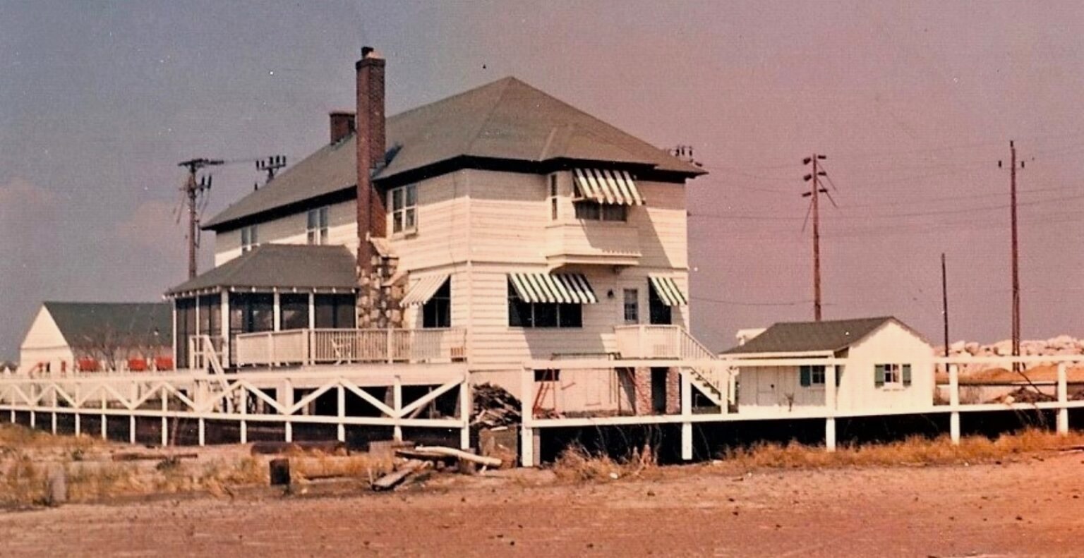 A color photograph ca. 1962 of the Sandlass family house at Highland Beach — three-story clapboard with green-and-white striped awnings, screened porch, stone chimney, picket fence in the foreground