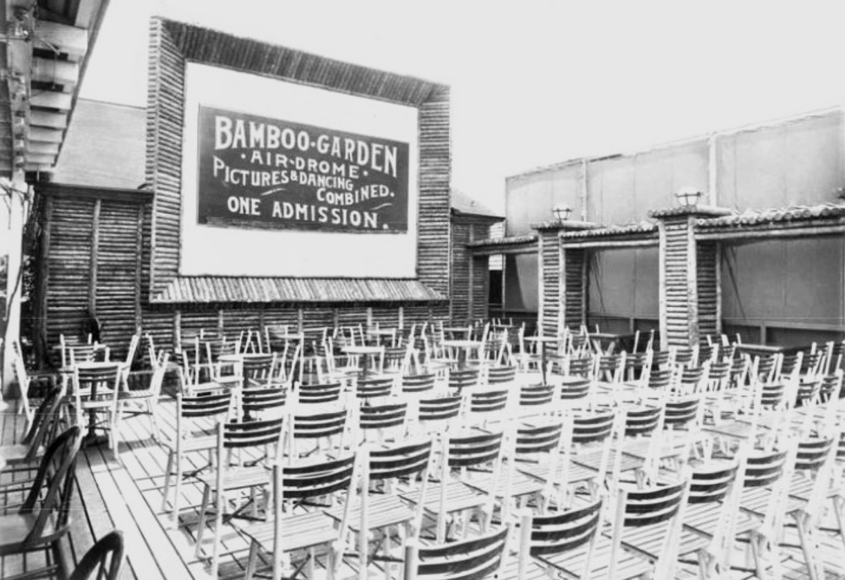 The interior of the Bamboo Garden Airdrome outdoor movie theater at Highland Beach, ca. 1922 — a bamboo-framed projection screen with signage reading BAMBOO-GARDEN AIR-DROME PICTURES & DANCING COMBINED ONE ADMISSION, rows of wicker folding chairs on a wooden deck