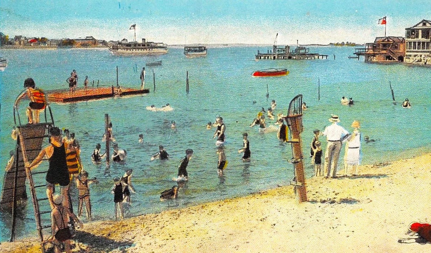A hand-colored postcard ca. 1928 of the Highland Beach swimming area — bathers on and around a floating raft, a diving platform on the sand, a small steamer and speedboats in the bay, the Sandlass Pavilion at right