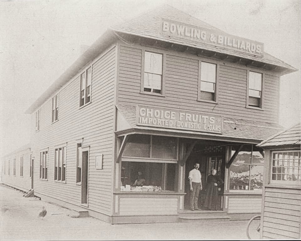 A sepia photograph ca. 1900 of a two-story wooden corner storefront at Highland Beach — signs reading BOWLING & BILLIARDS above and CHOICE FRUITS · IMPORTED & DOMESTIC CIGARS below the awning, a shopkeeper and a woman at the entrance