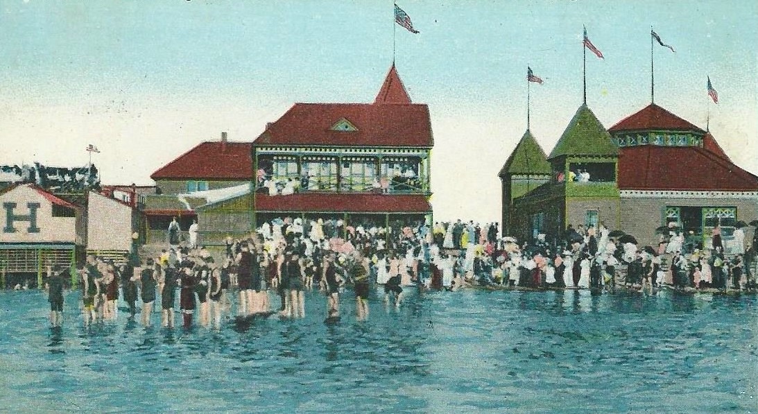 A hand-colored postcard ca. 1912 of Highland Beach bathing — several hundred bathers in Edwardian bathing costume in the shallow water, the Sandlass Pavilion complex with three American flags behind, striped bathhouses at left