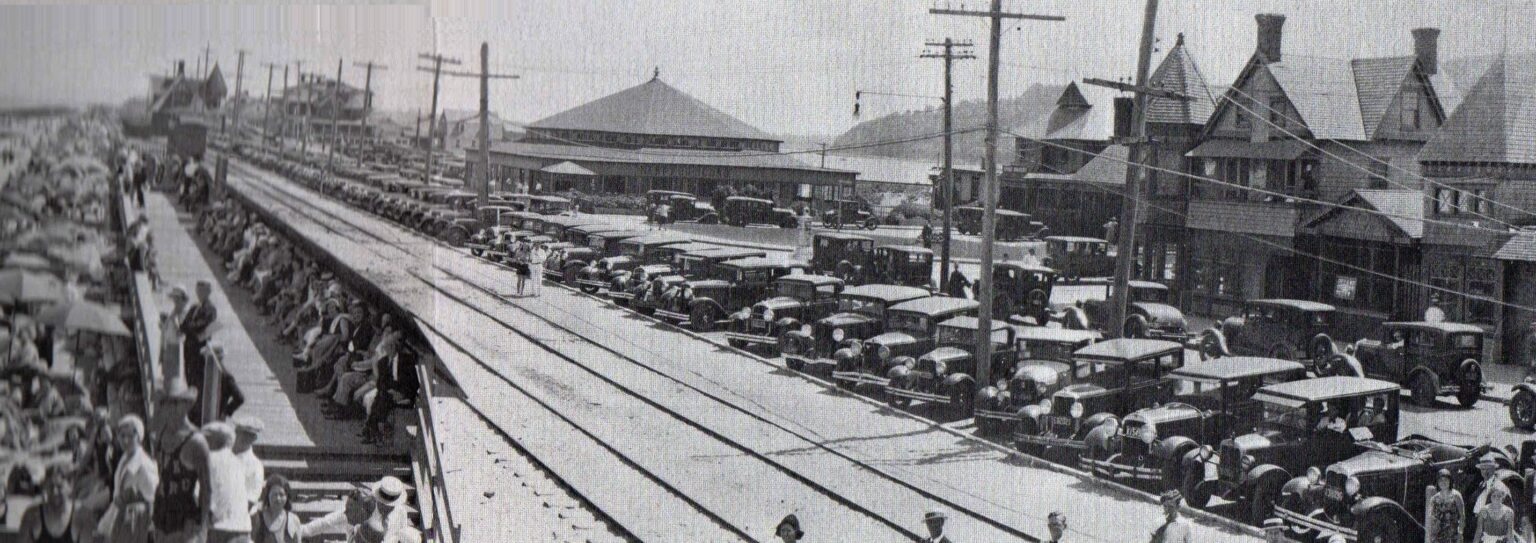 The Highland Beach railroad station platform on a summer Saturday ca. 1928 — a full platform of day-trippers at left, a row of 1920s automobiles at right, the hip-roofed station building, the Sandlass Pavilion behind