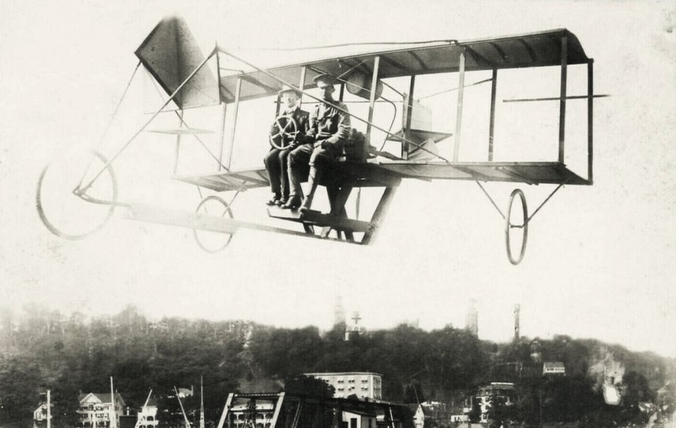 An early pusher biplane ca. 1911 in flight — pilot and passenger seated on the leading edge of the lower wing, front canard elevator and rear pusher propeller — with the Highlands ridge in the background and cottages at the base of the ridge