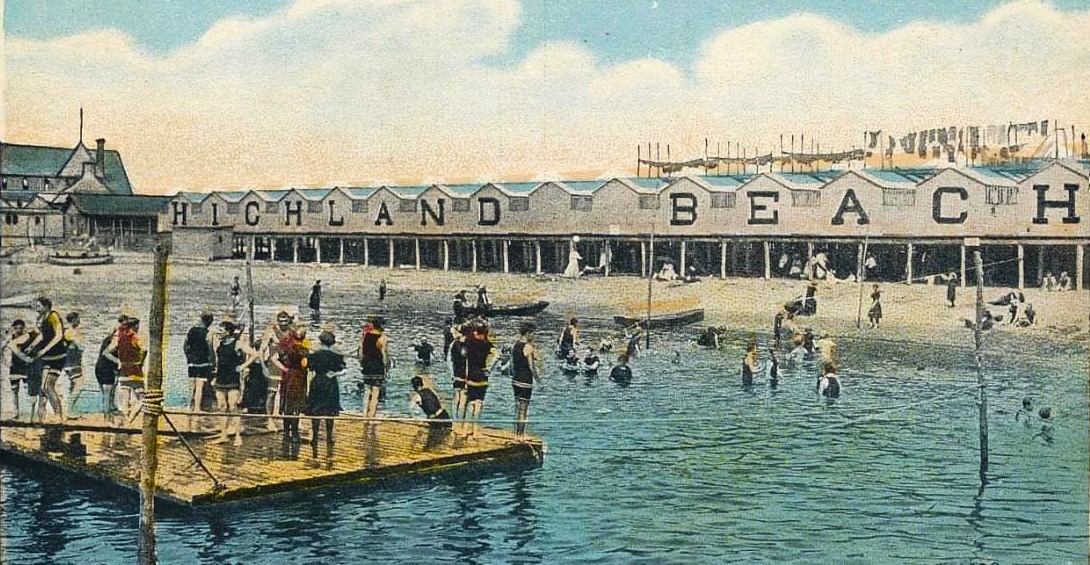 A hand-colored postcard ca. 1918 of the Highland Beach swimming area — a wooden diving platform at left with bathers standing on it, the HIGHLAND BEACH bathhouse row on pilings behind, swimmers in colored caps and striped suits in the water