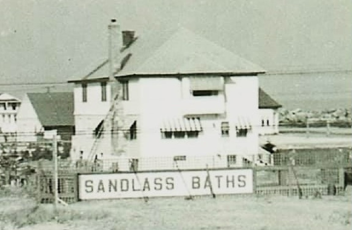 The Sandlass family house at Highland Beach ca. 1935 — a two-story stucco house with hipped roof and chimney, wooden bathhouses at right, a large white billboard-sized sign reading SANDLASS BATHS in the foreground