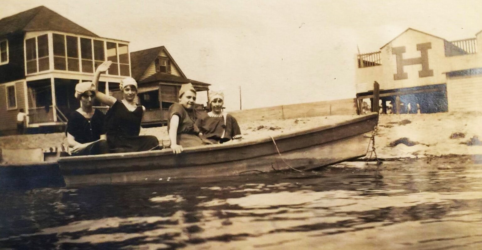 A sepia photograph ca. 1918 of four women in period bathing costume and white swim caps posing in a wooden rowboat on the sand at Highland Beach — a bathhouse with an HB monogram at right, a screened-porch cottage at left