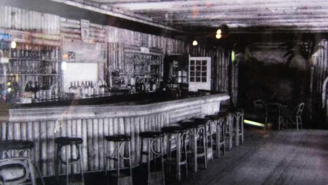 A sepia interior photograph ca. 1935 of the Bamboo Room bar at Highland Beach — a curved bar counter entirely faced with bamboo poles, mirrored bottle racks behind, Bentwood stools at the counter, and small wooden tables at right