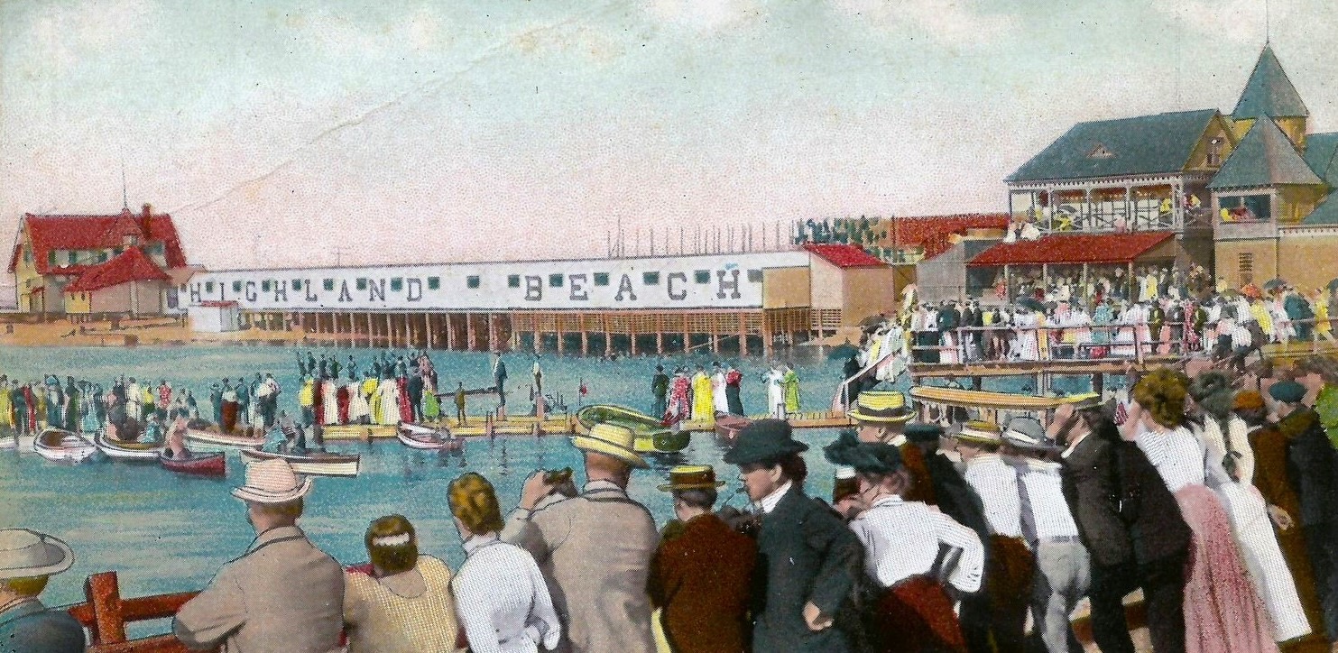A hand-colored postcard ca. 1908 of a regatta afternoon at Highland Beach — a large crowd along the shore, the HIGHLAND BEACH bathhouse row across the middle with painted lettering, the Sandlass Pavilion at right with crowds on both porches, a red-roofed cottage at far left