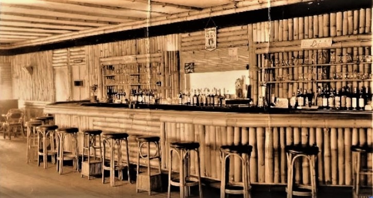 A sepia photograph ca. 1935 of the Bamboo Room bar at Highland Beach from a wider angle — the full run of the bamboo-clad counter, Bentwood stools, three rows of bottles on the back bar, walls and ceiling faced with vertical bamboo poles