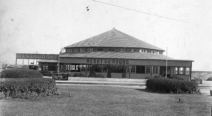 A sepia photograph ca. 1908 of the Merry-Go-Round pavilion at Highland Beach — a square wooden hip-roofed building with MERRY GO-ROUND painted across its facade, a RESTAURANT sign at left, ornamental hedges in the foreground