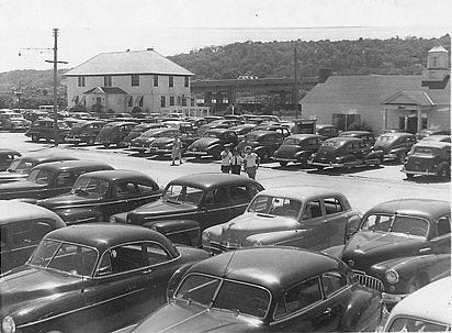 A sepia photograph ca. 1946 of the Sandlass Beach Club parking lot — postwar American sedans in the foreground, dozens more behind, the family house / future Building 600 in the middle distance, the white bath-house office at right, the Highlands ridge behind