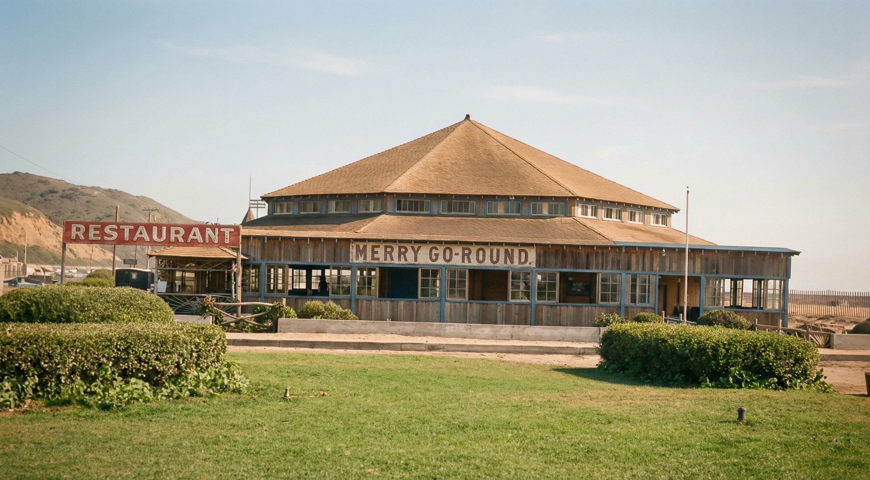 A color photograph ca. 1955 of the Merry-Go-Round pavilion at Highland Beach — a square wooden hip-roofed building with MERRY GO-ROUND signage and a RESTAURANT sign at left, blue window trim, mown grass in the foreground, the Highlands ridge at far left