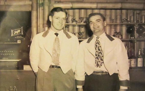 A sepia photograph ca. 1948 of two Bamboo Room bartenders behind the bar at Highland Beach — both in the short white bolero uniform jackets with brown collars and cuffs, brown trousers, patterned neckties, Rheingold signage and bamboo shelves behind