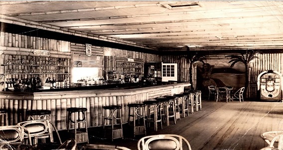 A sepia interior of the Bamboo Room bar at Highland Beach ca. 1948 — wider angle showing the full curve of the bamboo-clad bar, Bentwood stools, bottle shelves, a partial jukebox at right, the Hawaiian-mural walls in the corner