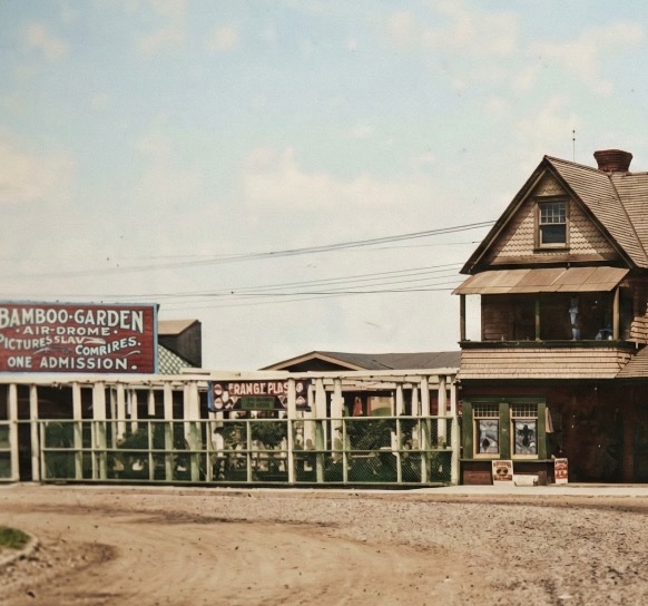 An AI-colorized detail crop ca. 1912 of the Sandlass entertainment complex plaza — a boardwalk gate at left, the BAMBOO-GARDEN AIR-DROME signboard clearly legible, ORANGE PHOSPHATE placard at right, the corner of the Victorian pavilion office