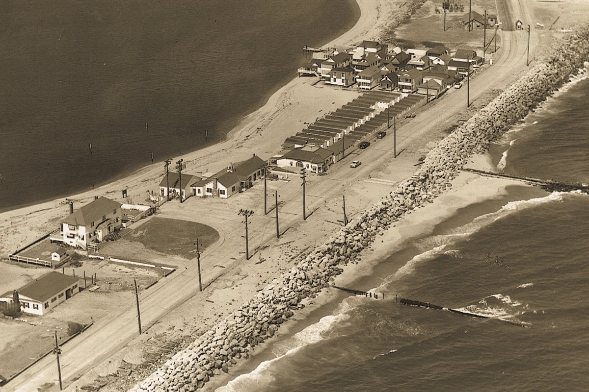 A sepia aerial photograph ca. 1955 of Highland Beach from the ocean side — the Shrewsbury bay at upper left, the cottage colony at upper right, the row of Sandlass bathhouses along Ocean Avenue at center, the family house at lower left, and a rock seawall with stone groins along the ocean front at bottom