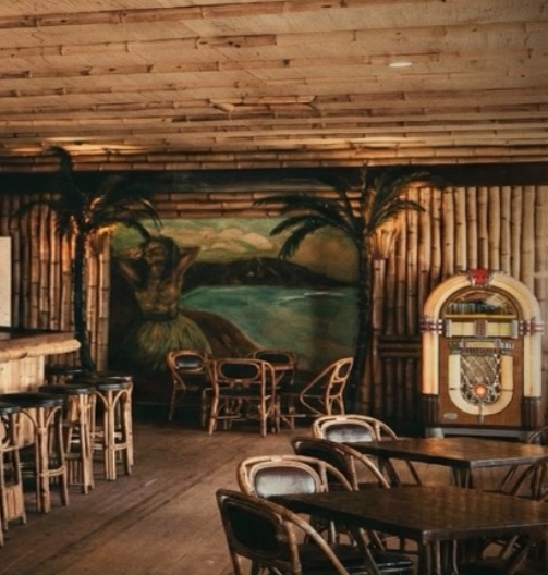 The east end of the Bamboo Room bar at Highland Beach ca. 1948 — a hula-dancer mural in oil paint on the wall framed by real bamboo poles, walls faced with vertical bamboo, Bentwood chairs and small wooden tables in the foreground, a lit Wurlitzer jukebox at far right