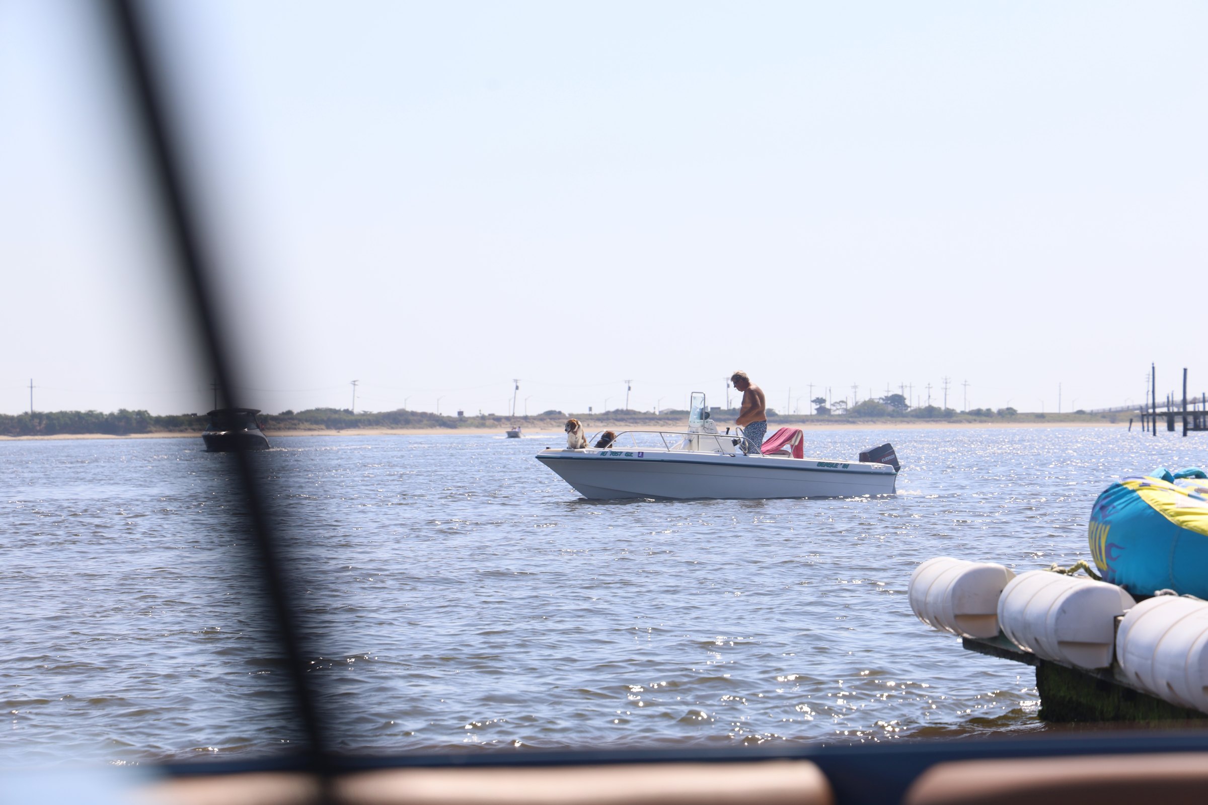 A small white center-console boat crossing calm bay water near a sandy spit, two people aboard with one standing at the console and a red-and-white diver's flag flying, framed past dark dock rigging in the foreground
