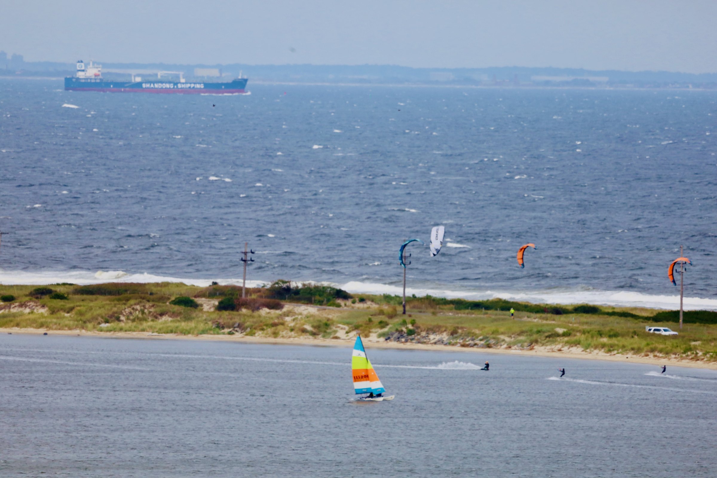 A bright windy day on the bay with whitecaps — a small catamaran under a rainbow-striped sail in the foreground, several kitesurfers with colorful kites off a barrier spit, and a container ship on the horizon