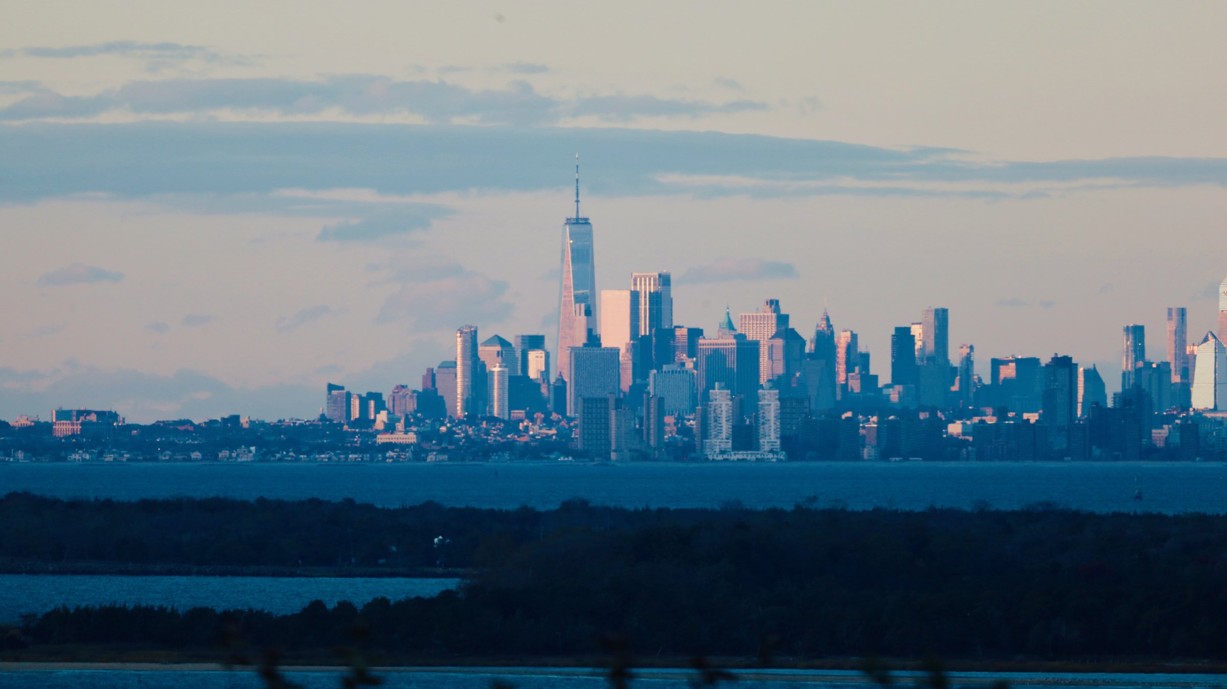 The Manhattan skyline lit against a deep blue twilight sky, seen across dark water from a height with a dark foreground ridge