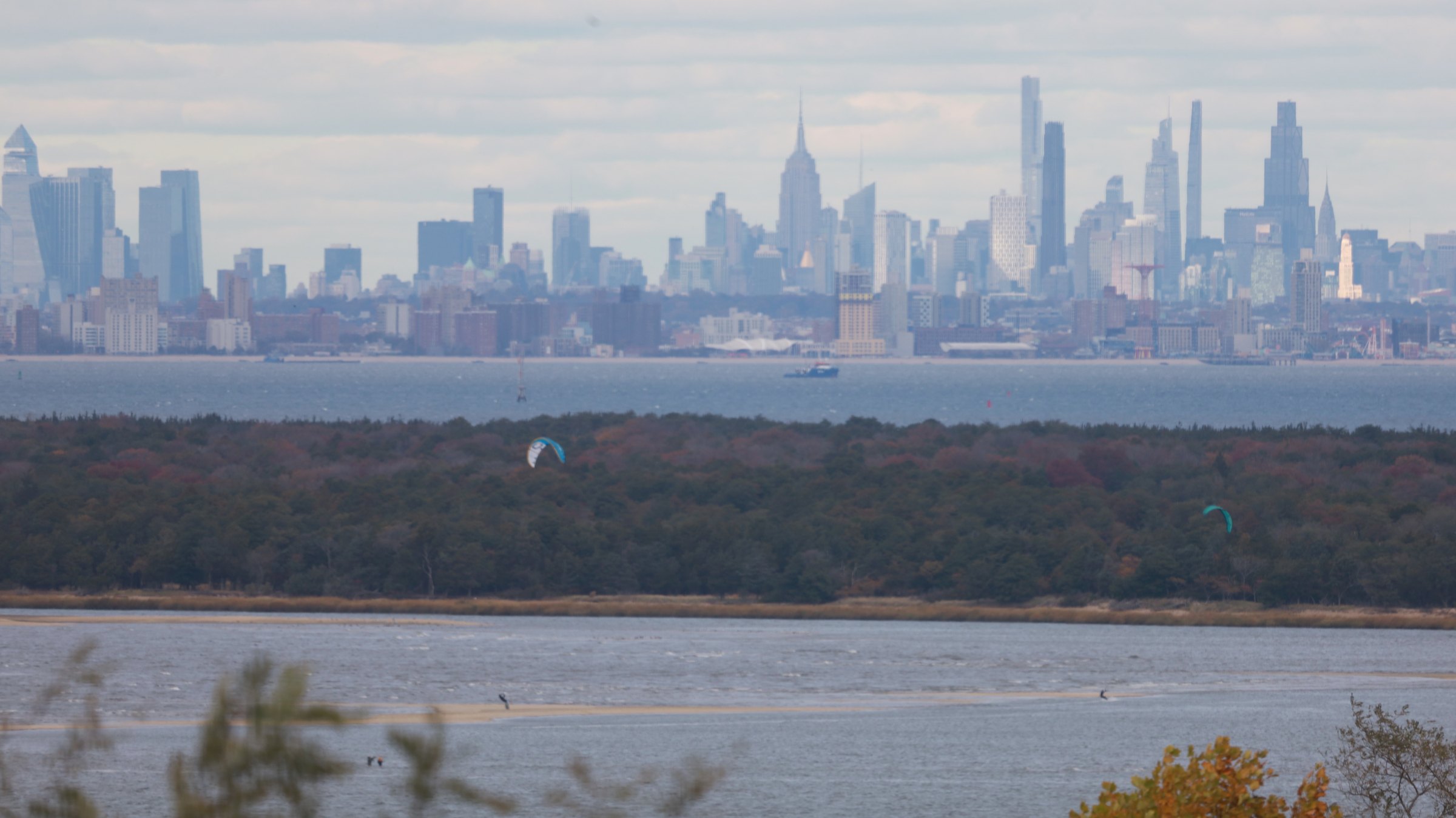 The Manhattan skyline low across blue bay water beyond a long low green barrier spit under a bright hazy sky