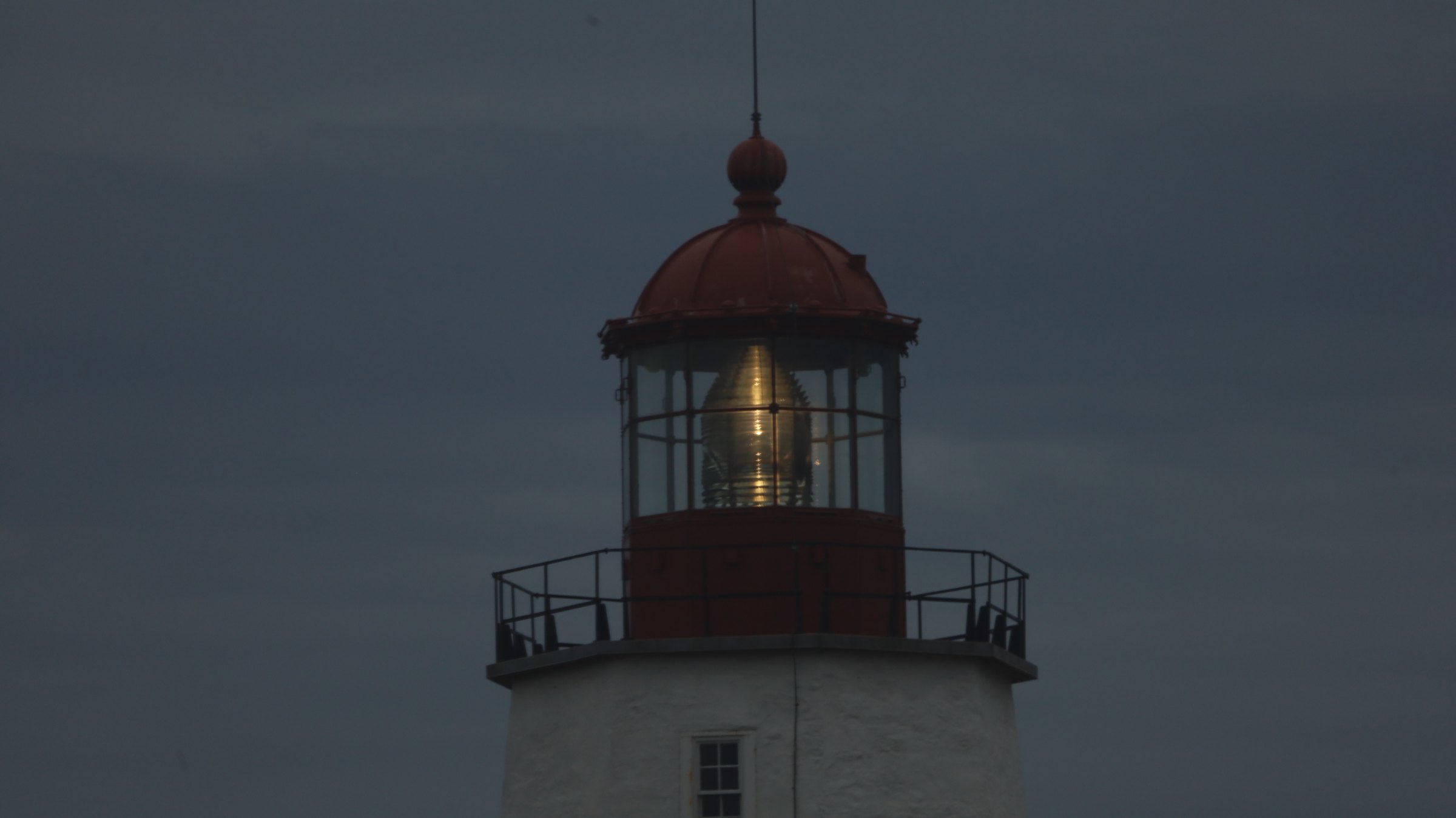 The lantern of the Sandy Hook Lighthouse close up at dusk — a red domed lantern room with a large lit Fresnel lens glowing behind the glass, a black gallery railing below