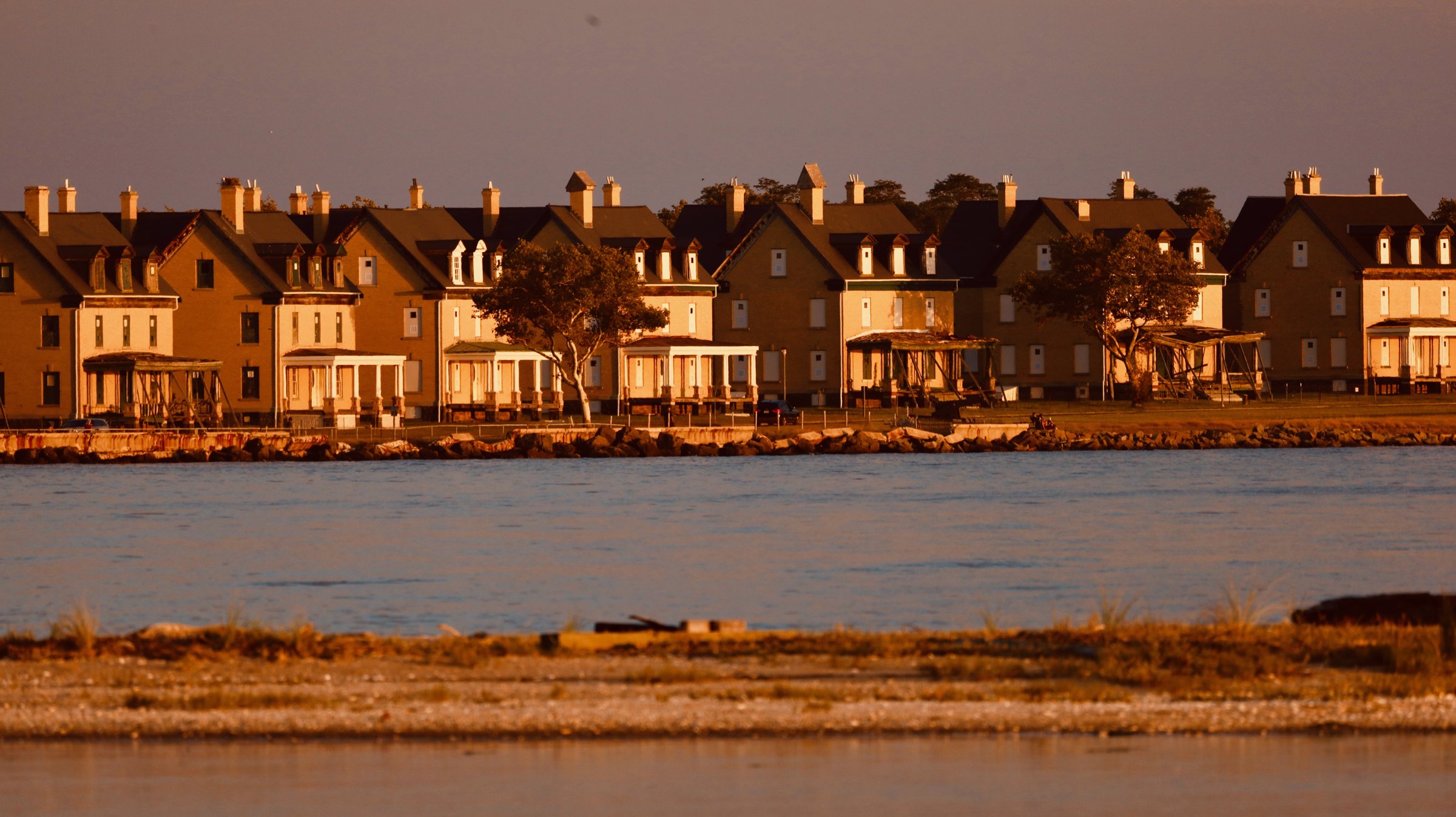 A long row of peaked-roof shorefront houses with porches across a strip of water, warm in low golden sunset light