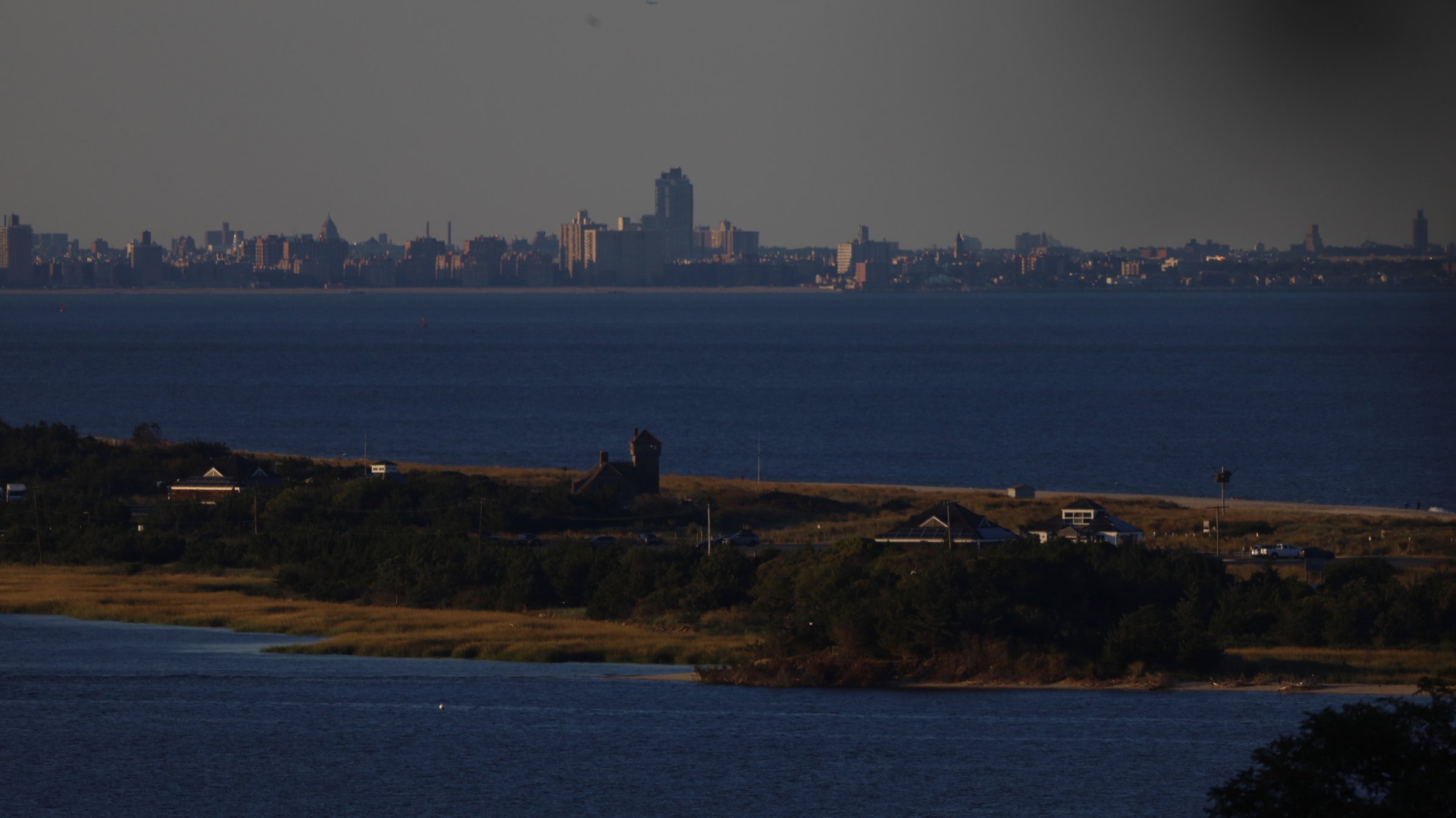 The distant Manhattan skyline low on the horizon across a wide bay at dusk, seen over a barrier spit and salt marsh from an elevated vantage, a single tower standing on the sand