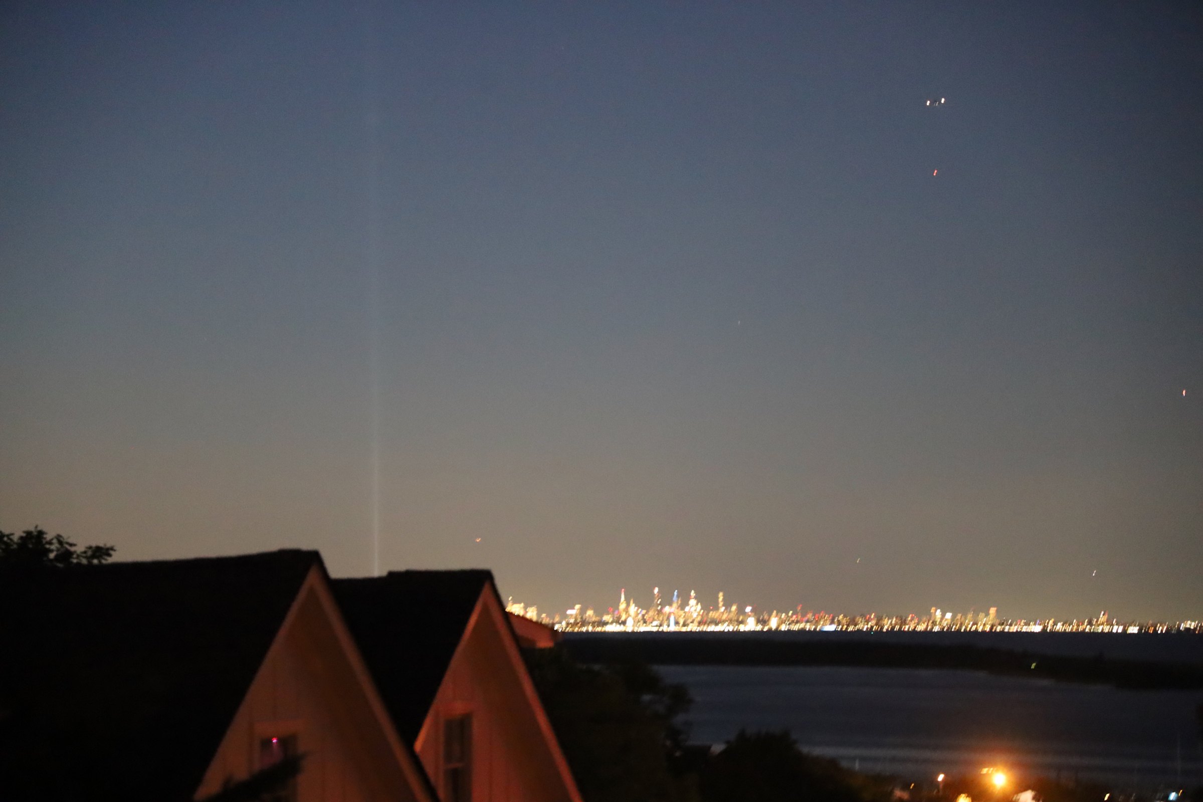 The Manhattan skyline glittering as a band of light low across a dark bay at night, seen over the peaked rooftops of shore houses, a single beam of light rising into a deep blue sky