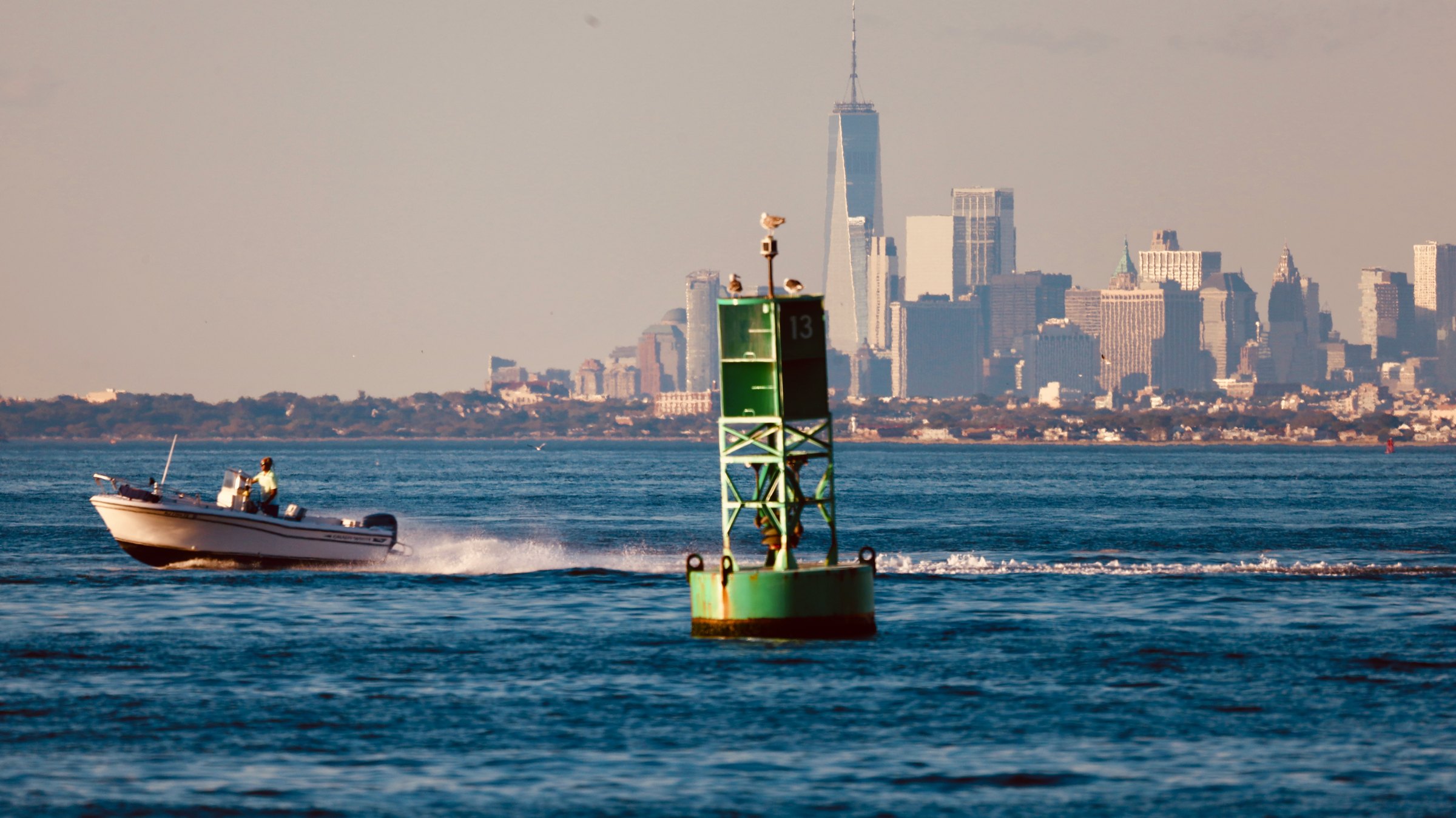 A small open boat passing a large green channel buoy on choppy bay water, the Manhattan skyline stretched low across the horizon behind