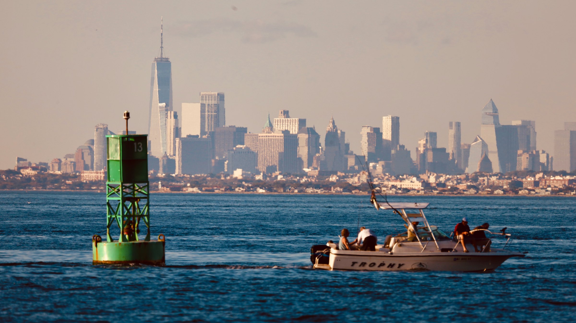 A fishing boat with figures aboard passing a green channel buoy on the bay, the Manhattan skyline low across the horizon behind