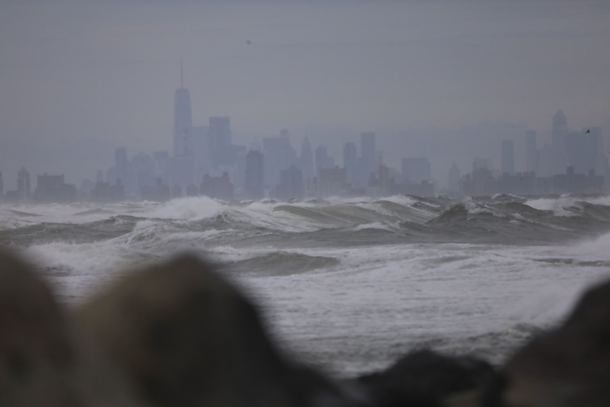 Rough grey ocean surf with breaking whitecaps under an overcast sky, the Manhattan skyline faint and hazy on the horizon behind