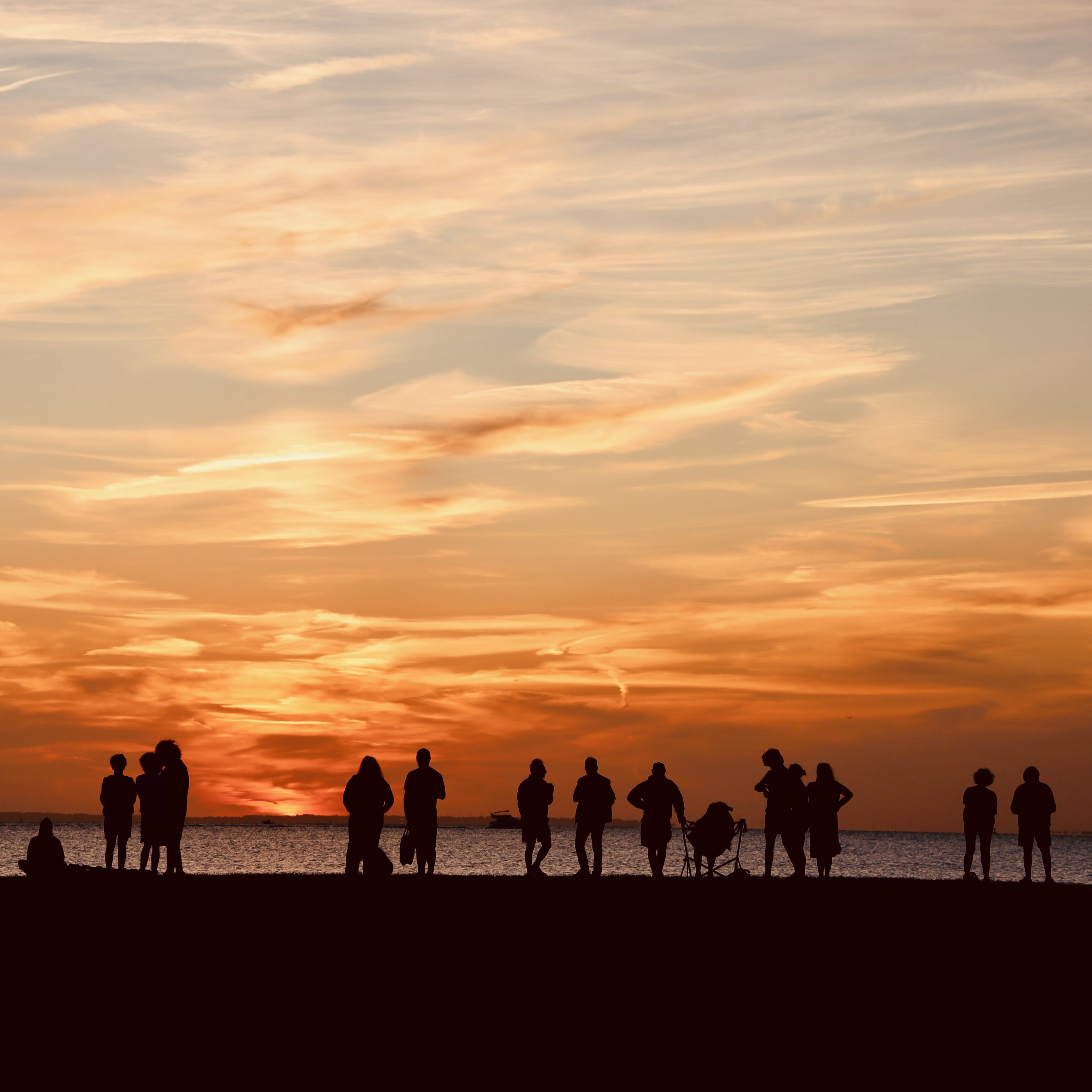 A row of people and beach chairs silhouetted on the sand against a vivid orange-and-grey sunset sky over the water