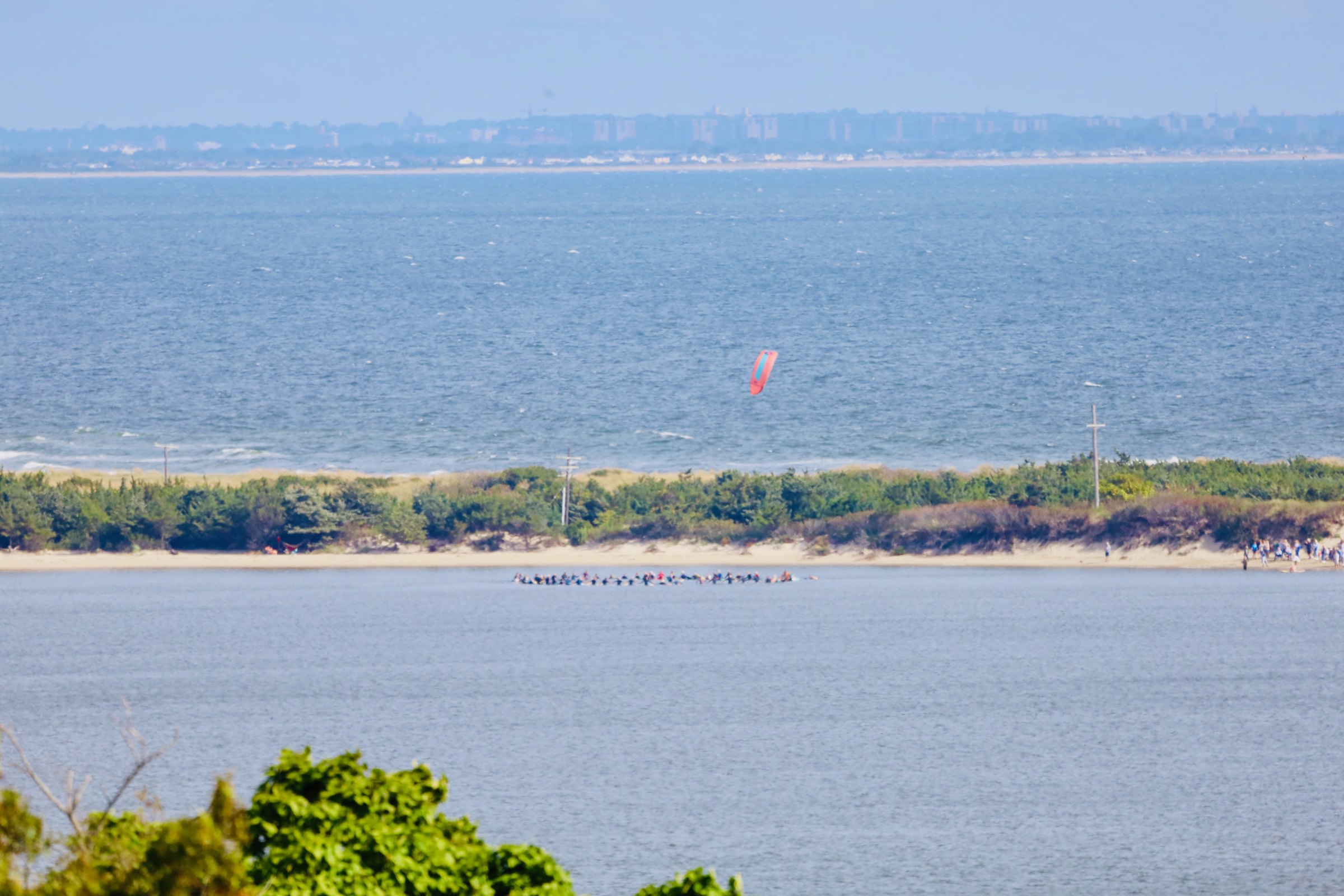 A wide view over blue bay water to a low sandy barrier spit, a kitesurfer on the water and a hazy far shore under a bright sky