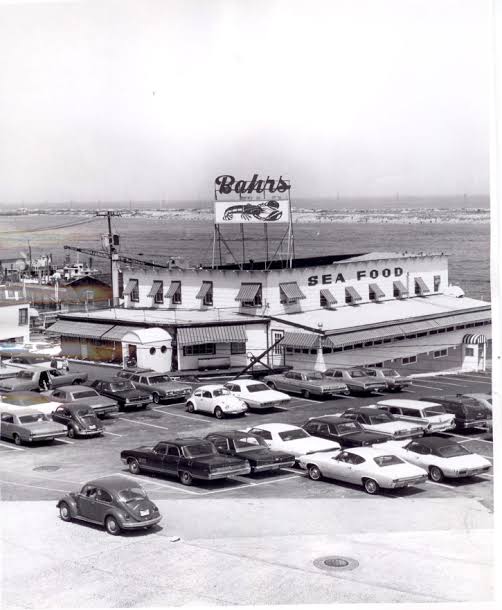 A black-and-white photograph of Bahrs Landing from the Bud and Peg era — the rooftop Bahrs lobster sign and SEA FOOD lettering over the restaurant, a full parking lot of period cars in the foreground, Sandy Hook visible across the water.