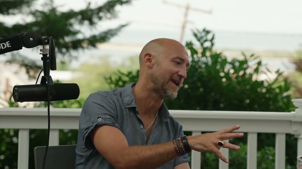 Anthony Setaro mid-sentence at a microphone on a porch, one hand raised, green trees and a strip of bay behind him.
